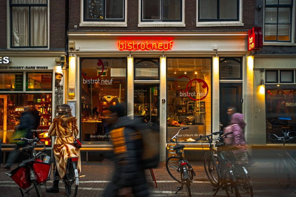 Exterior view of Soupenzo, a popular soup shop, showcasing its warm, inviting atmosphere through large glass windows. The shop is located on a bike-lined street in the city, with patrons enjoying their meals inside on a chilly evening.